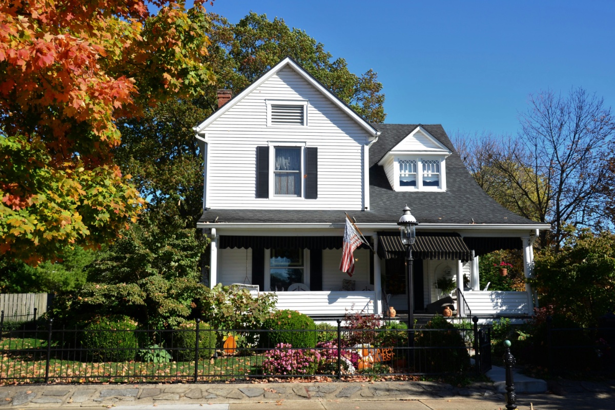 White two-story house with black shutters and awnings, surrounded by green shrubs and trees with some orange autumn leaves, a black metal fence in the foreground, and an American flag hanging on the porch. The appraisal came in low.