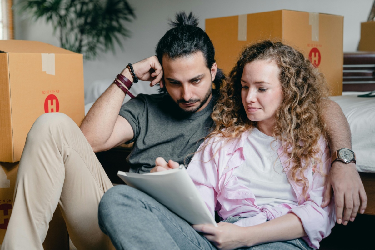 A couple sits on the floor amidst moving boxes, intently reviewing paperwork related to a down payment gift for their new home.