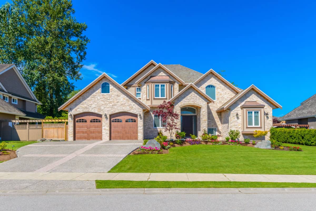  A grand, light-colored stone house with multiple gables and large windows stands under a clear blue sky. Two wooden garage doors are visible on the left, leading to a curved driveway. The front yard features a meticulously manicured green lawn, colorful flower beds, and a small, red-leafed tree near the entrance. A tall green tree is visible on the left, and a paved street runs across the foreground. This image conveys the idea of a dream home or a luxury residence. minimum down payment for house first time buyer