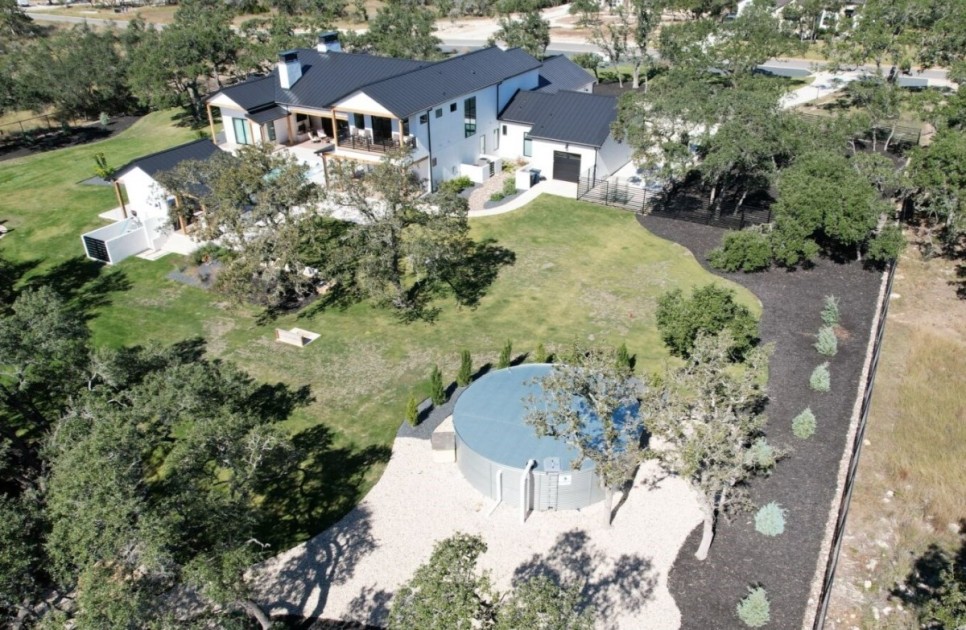an angled birds eye view of a property with a white barn house, large water harvesting tank, and lush green grass