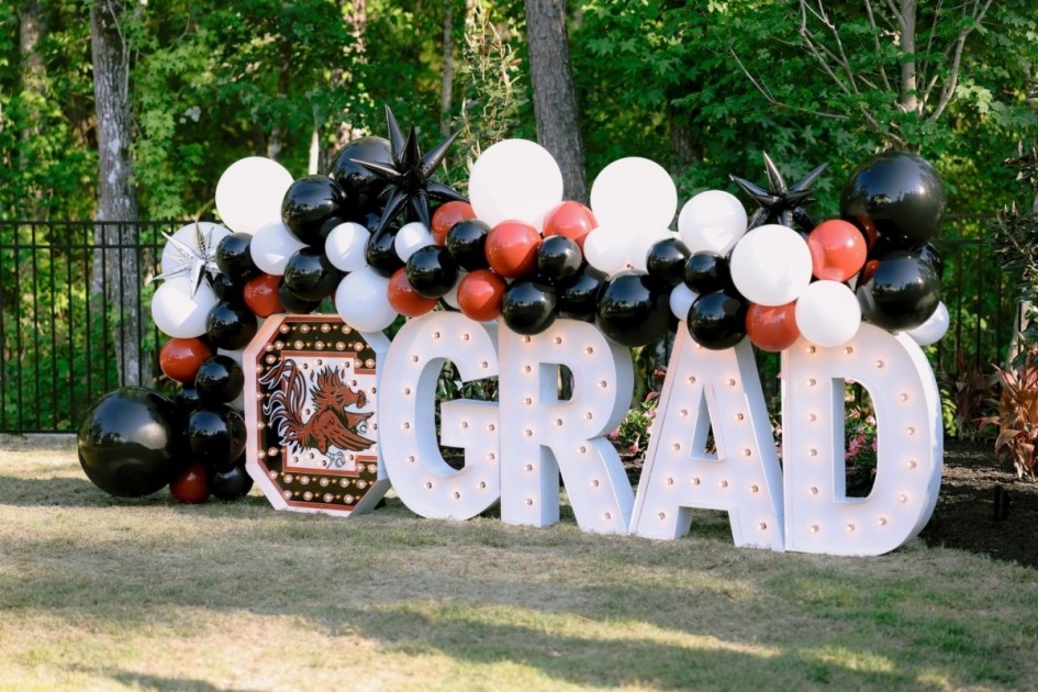 an LED sign reading "GRAD" sits outside decorated with an LED of the school's mascot and school colored balloons at a home graduation party