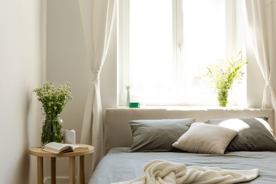 A linen bed next to a bright window with flowers on the bedside table