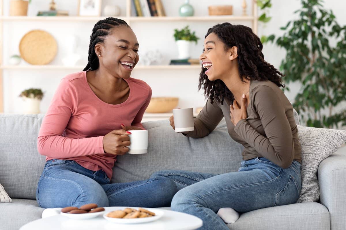 Two friends sit on a couch with coffee and cookies discussing how to buy a house with a friend.