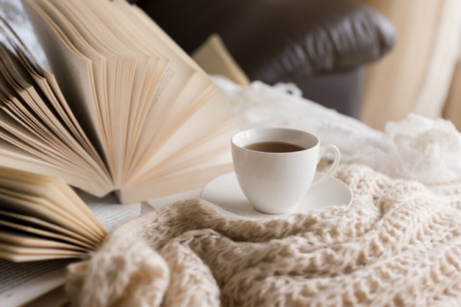 A cup of warm tea sits on a cozy blanket next to an open book