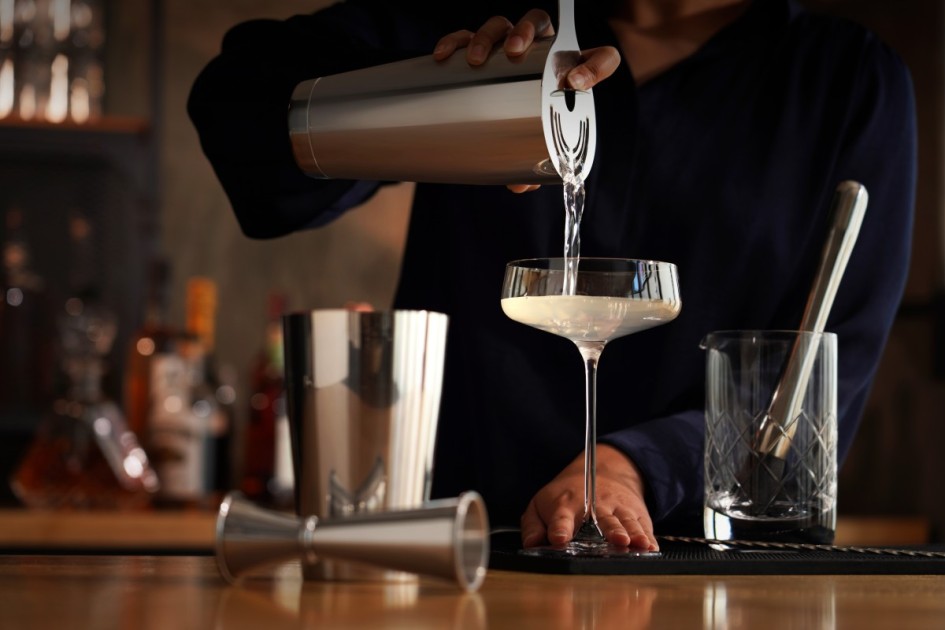 a person wearing a dark blue long sleeve pours a drink into a glass from a silver shaker with other bartending tools on the wooden home dry bar