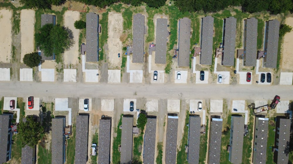 Windgate Park neighborhood features rows of elongated prefab dwellings flanking central gravel roadway. Series showcases uniform concrete parking slabs linking housing units in New Braunfels, TX