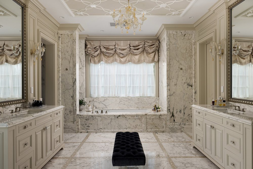 Opulent primary bathroom with extensive marble finishes, dual vanities, a deep soaking tub under a draped window, and a central tufted bench.