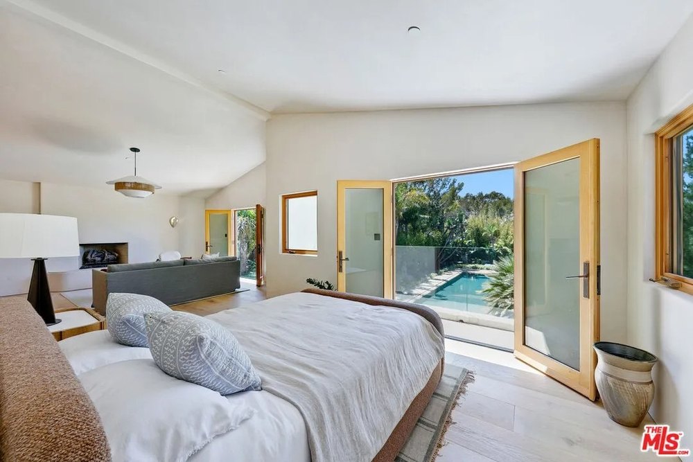 A bedroom with a king-sized bed and blue patterned pillows has open glass doors that lead directly to a private balcony overlooking the pool.
