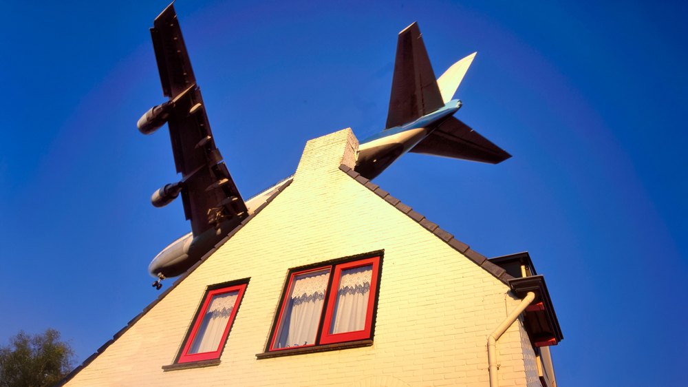 A plane in a blue sky flying over a white home with red trim windows