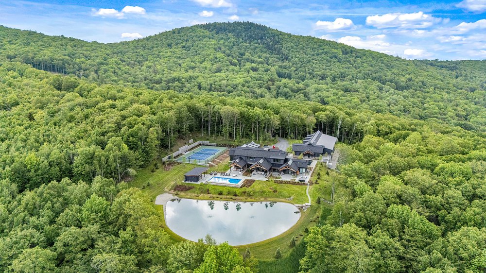 High-altitude drone shot of a modern estate tucked into a vast green mountain forest with a private pond.