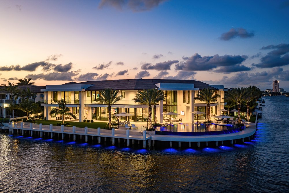 Mansion Sunset Elevation: The water-facing facade of a modern luxury estate glows with warm interior lighting against a dramatic sunset sky, accented by blue seawall lights.