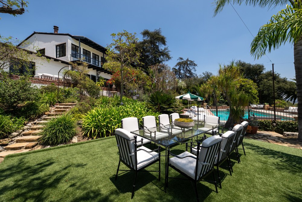 Outdoor glass dining table and chairs on a manicured green lawn behind a luxury home.