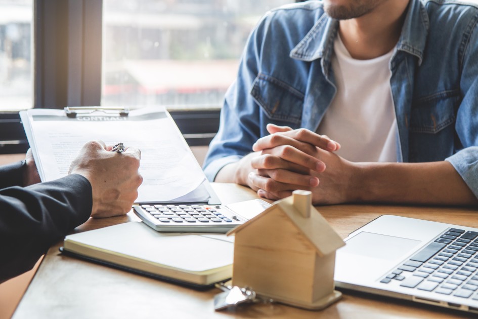 A person sits across a desk from someone explaining a contract. There is an open laptop, a small wooden house figurine, and a calculator on the desk between them. The person is considering buying a house with an LLC. 