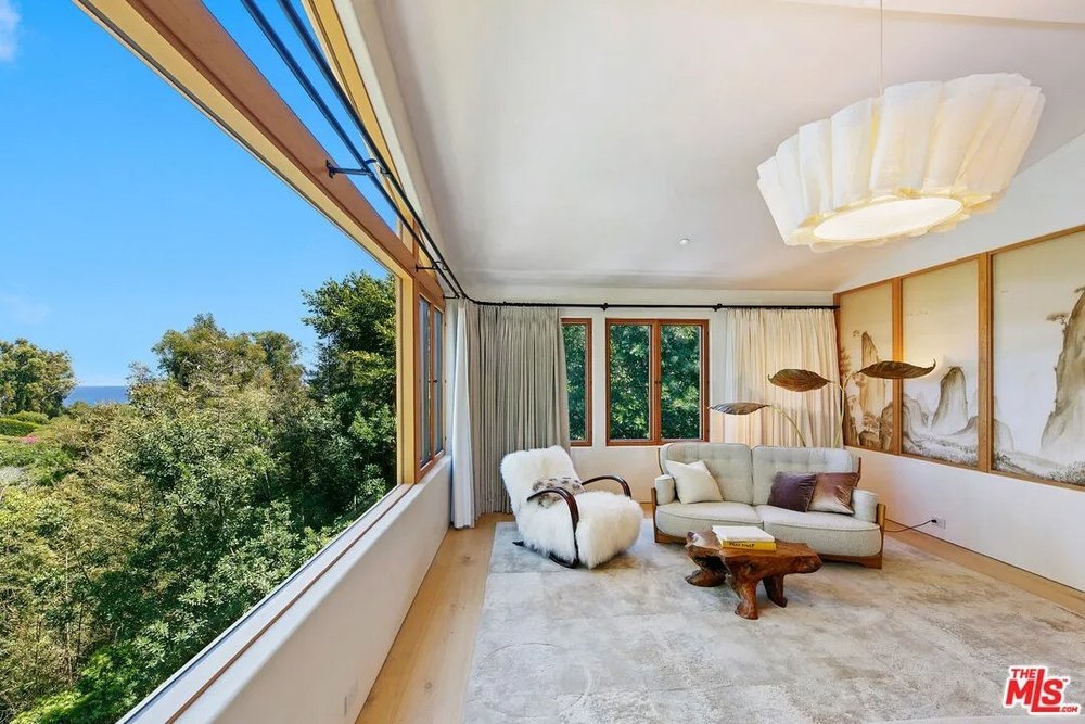 A small lounge area features a white fur armchair, a grey sofa, and a large panoramic window overlooking a dense forest.