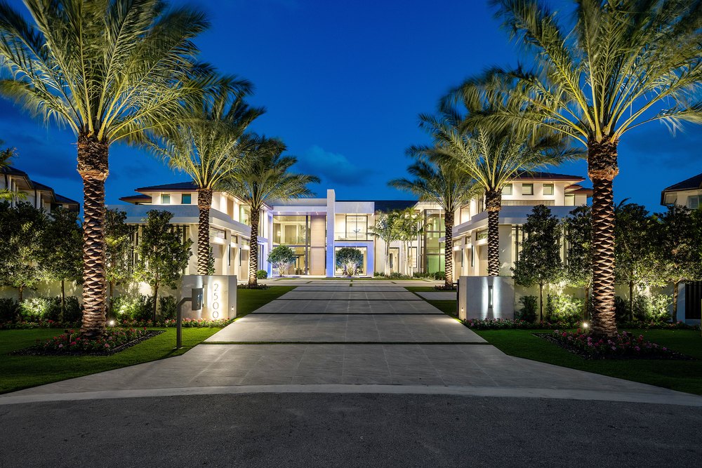 Dusk Lighting: Dramatic architectural uplighting illuminates the estate's palm trees, manicured landscaping, and glass facade against a deep twilight sky.