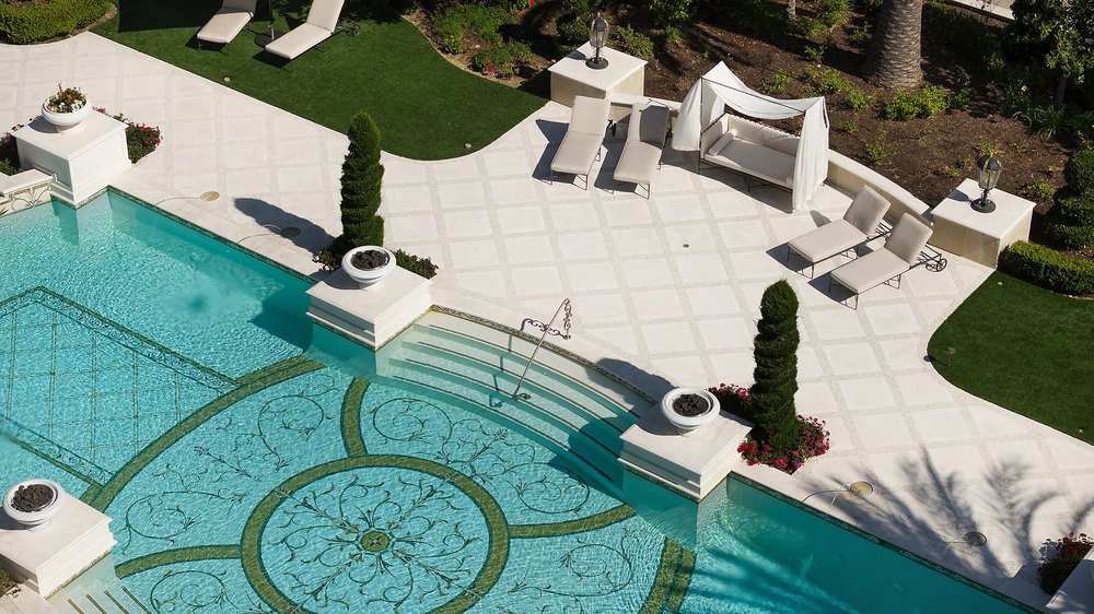 Overhead view of a sun-drenched pool deck featuring patterned tilework, lounge chairs, a cabana bed, and sculpted topiaries.