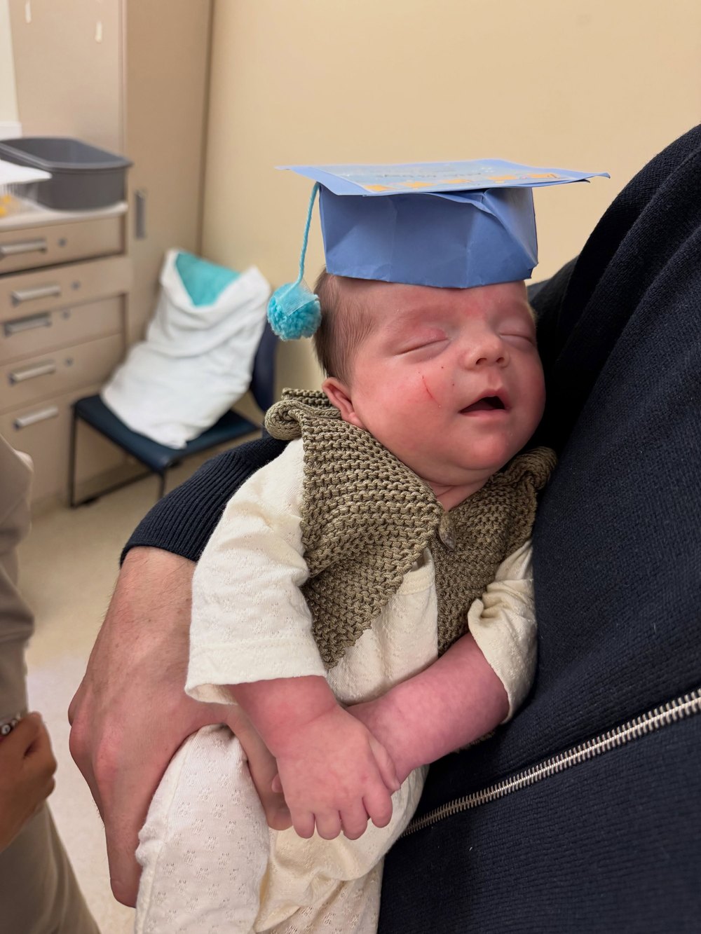 Close-up of a father cradling his sleeping newborn baby who is wearing a tiny blue paper graduation cap with a light blue tassel.