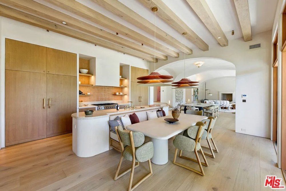 A view of a modern kitchen with natural wood cabinets, a plaster-finished island, and an integrated, curved breakfast banquette with green upholstered chairs.