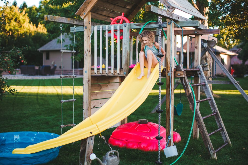 Little girl sits at the top of a backyard play structure as a hose she had rigged up sprays onto the slide as she gets ready to slide down.