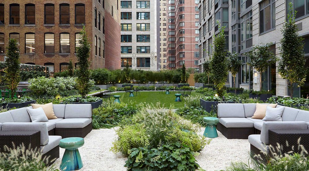 Landscaped rooftop garden courtyard with crushed gravel pathways, grey outdoor sectional couches, green ceramic stool tables, manicured lawn, and surrounding city buildings.