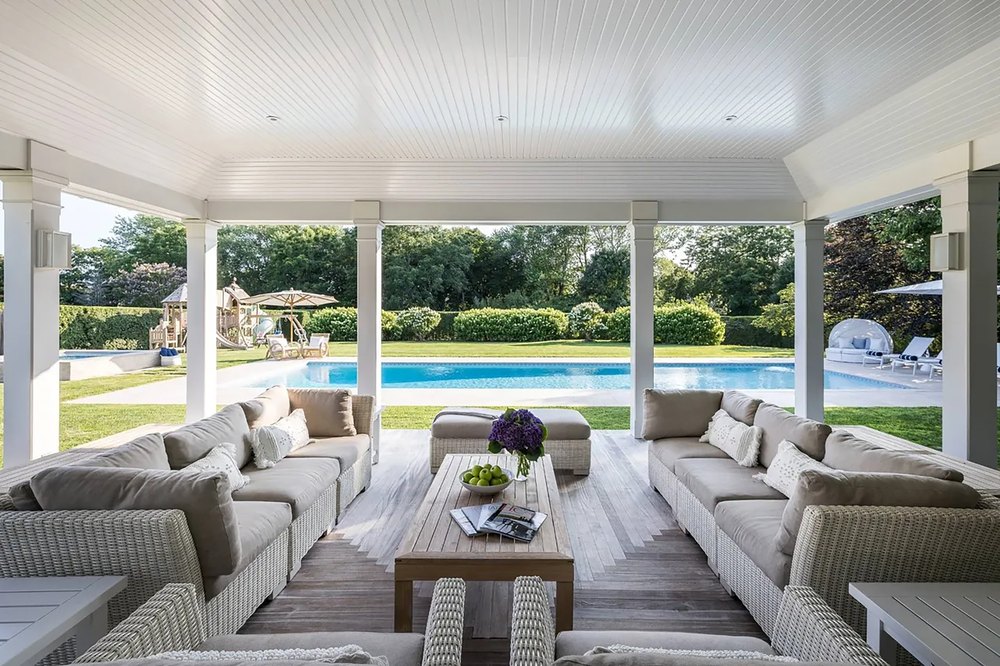 Covered patio lounge with white wicker sectional seating and a wooden coffee table overlooking a swimming pool and vast green yard.
