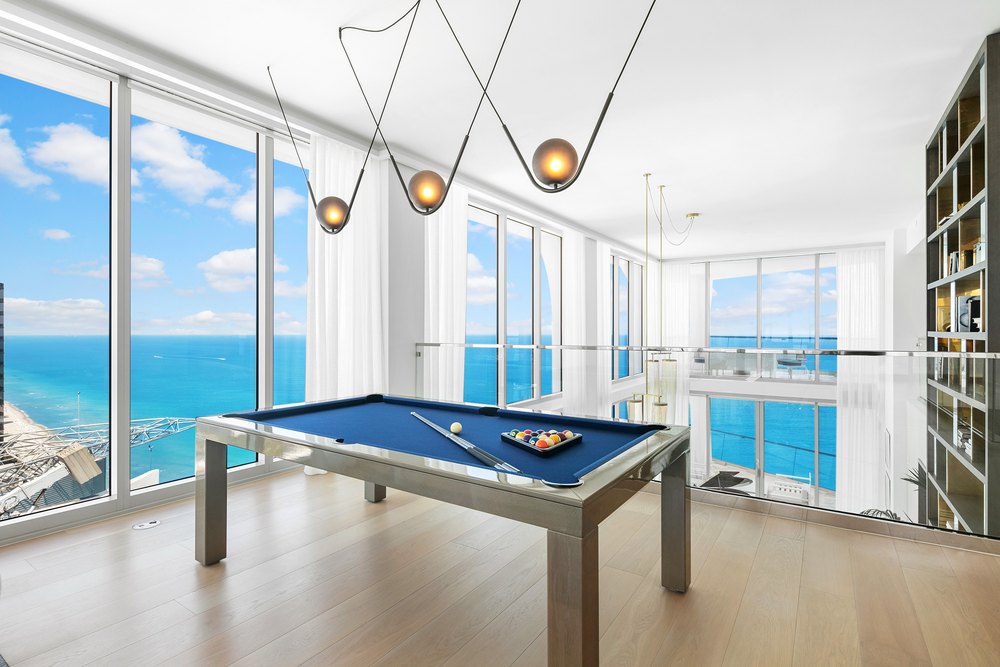 Penthouse mezzanine lounge featuring a sleek billiards table, glass guardrails, modern pendant light fixtures, and expansive ocean vistas.