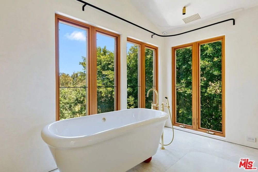 A white porcelain soaking tub with gold fixtures is positioned in a corner surrounded by three large windows looking out at green trees.