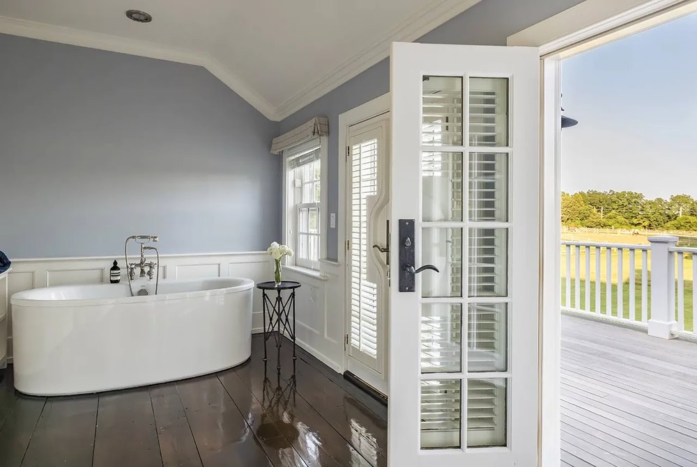 Primary bathroom corner featuring dark wood floors, a freestanding white oval soaking tub, white wainscoting, light grey walls, and a glass door opening to a sunlit deck.