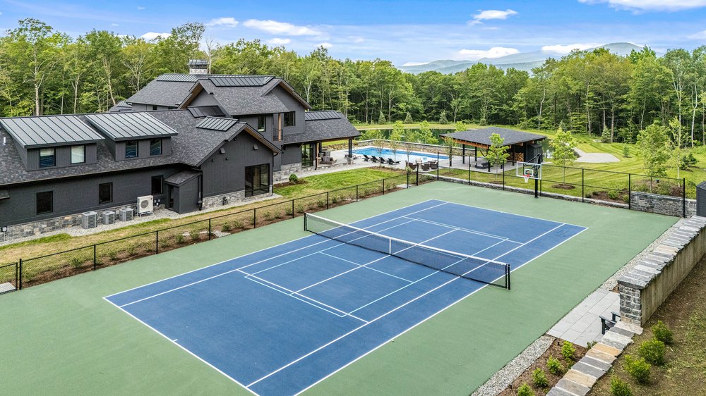 Aerial view of a full-sized outdoor tennis court and basketball hoop on a mountain estate property.