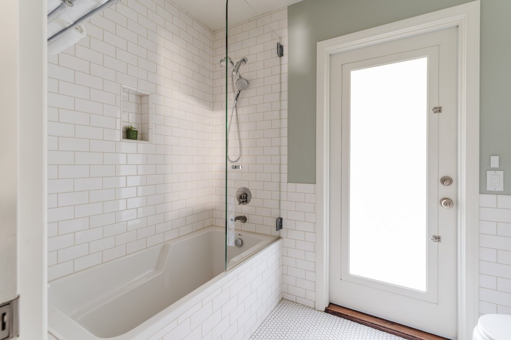 A bright bathroom featuring a white subway-tiled bathtub and shower combination with a glass partition.
