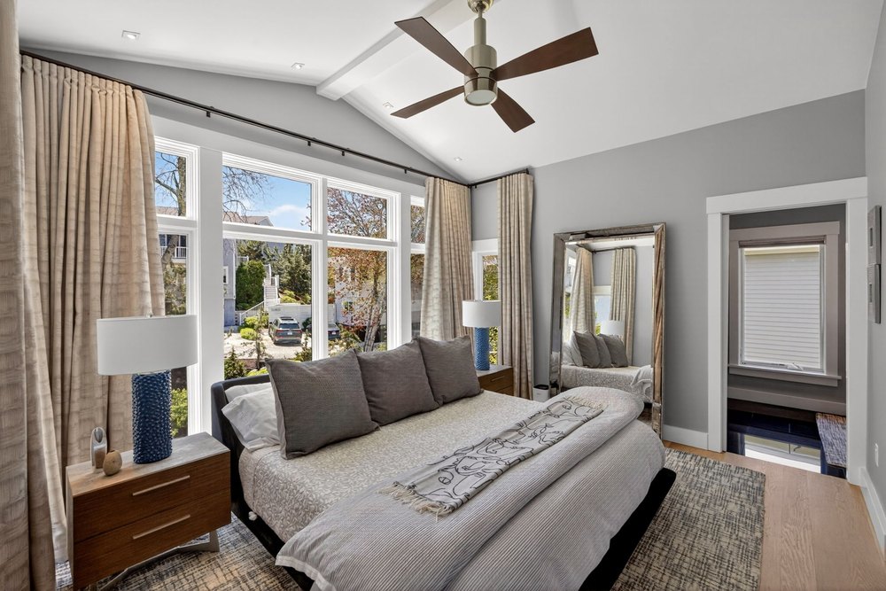Spacious primary bedroom with vaulted ceiling, floor-to-ceiling drapes, plush king bed, full-length mirror, and large windows.