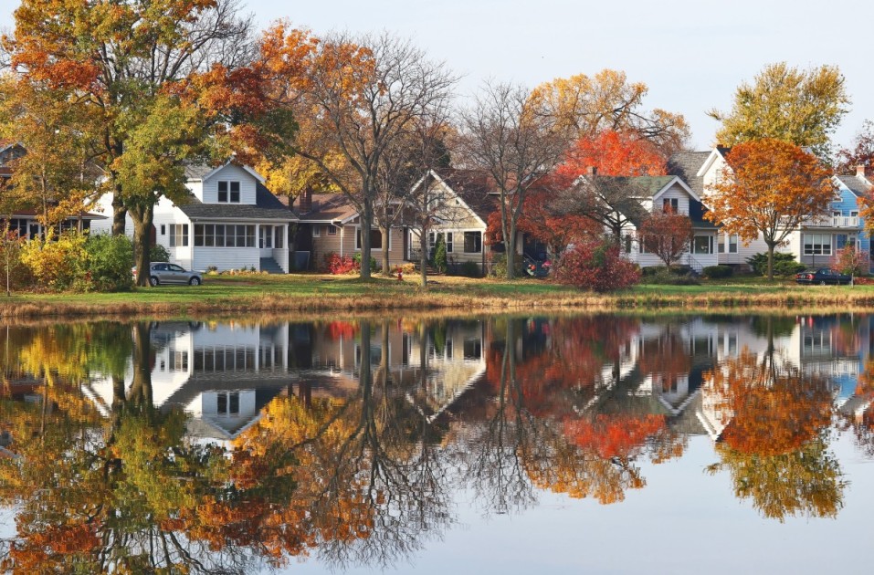 Houses and autumn trees reflect in the water from a lake, which might be a perk of buying a house in a flood zone. 