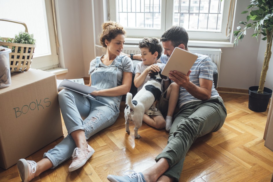 A family of three with a dog sit on the floor among moving boxes as they prepare to move to the home they are buying out of state. 