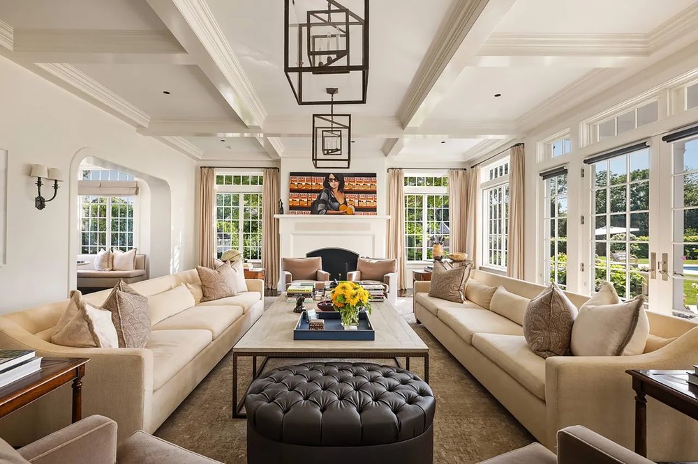 Formal living room with coffered ceilings, dual cream sofas, a tufted leather ottoman, a fireplace, geometric lantern chandeliers, and tall windows.
