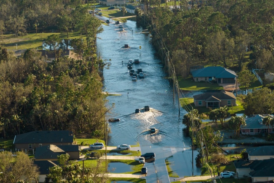 A flooded street with cars driving through the water. Many houses in this flood zone have water in their driveways or coming up to the house itself. 