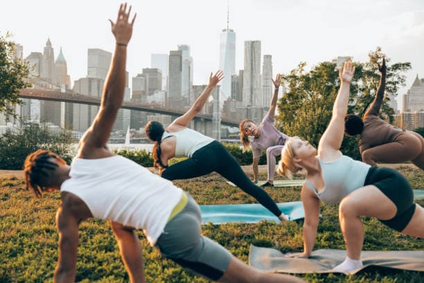 Group of people attending to a yoga class outdoors at sunset with New York cityscape on their background. They are meditating and relaxing.