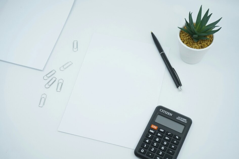 A calculator, pen, two pieces of paper, potted plant, and paperclips sit on a white table