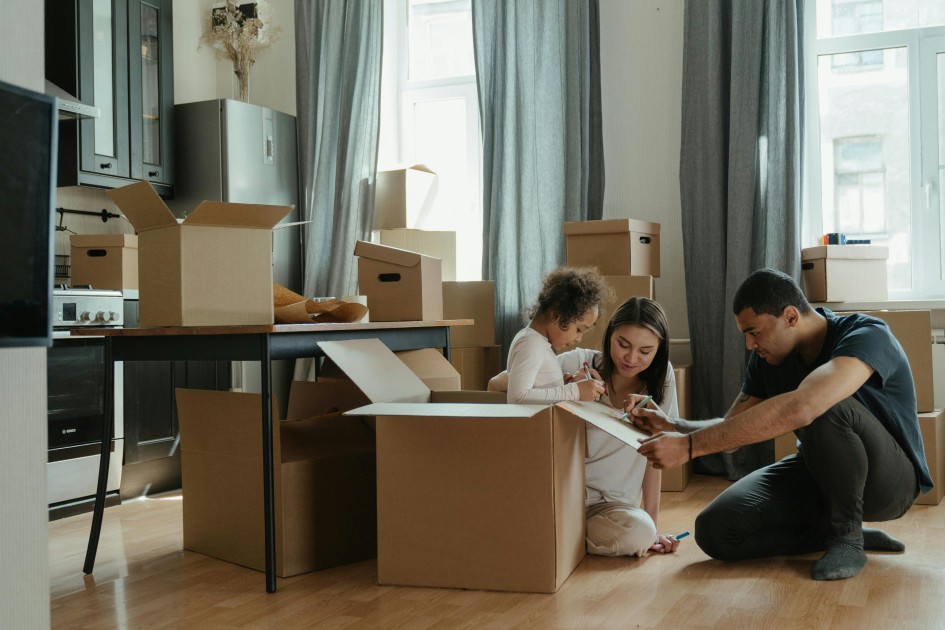 a couple sits on the floor of their home with a child surrounded by moving boxes after having decided on purchasing a new construction home