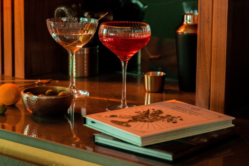 two coupe glasses with color cocktails sit in front of a shaker and next to a stack of books at a home dry bar