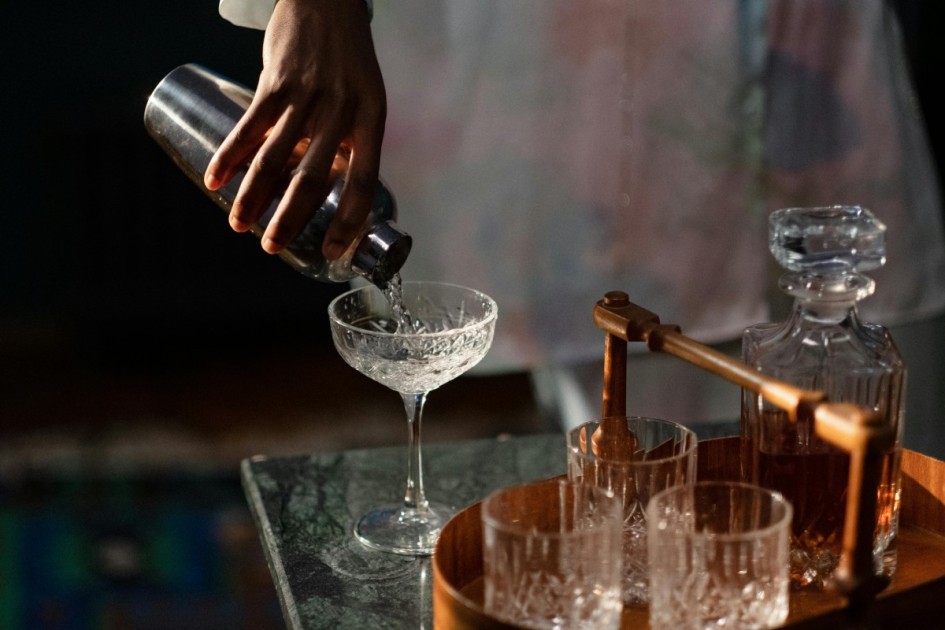 a hand pours a drink into a crystal glass from a silver shaker onto a the marble countertop of a home dry bar 