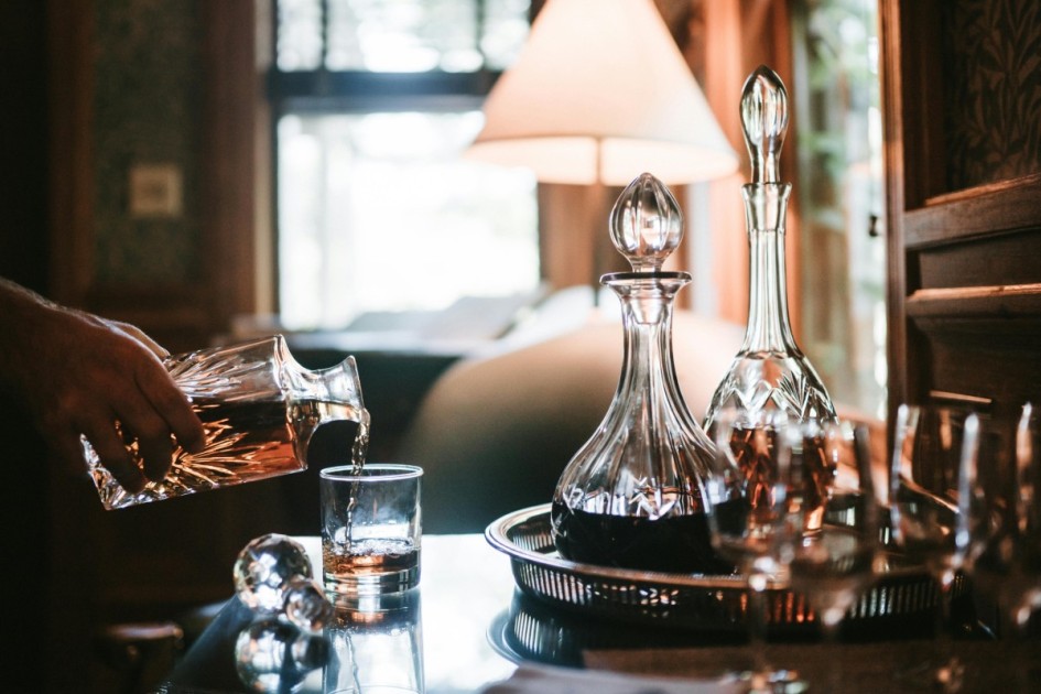 a hand pours whisky into a class from a decanter at a dark wooden home dry bar