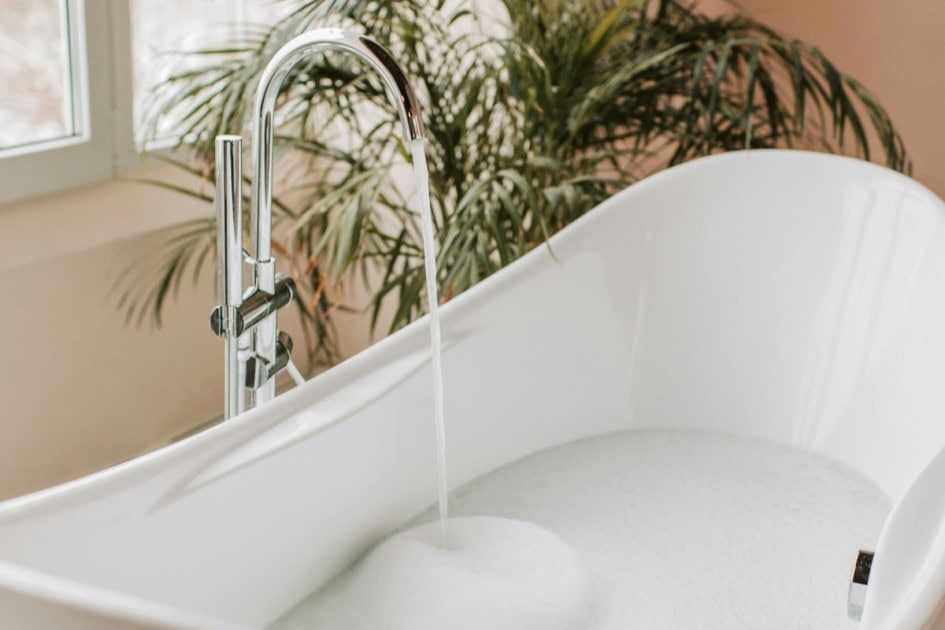 a chrome faucet fills up a large white tub with a leafy plant in the background begging the questions how to estimate utility costs when buying a house as answered in this Redfin article