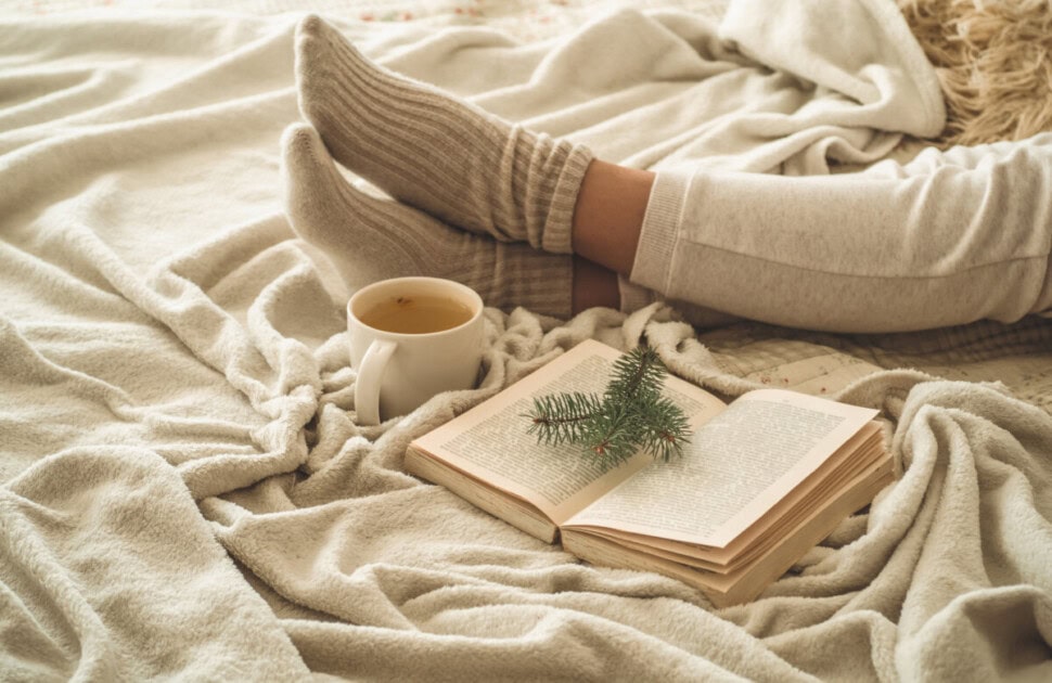 Feet with warm socks on them lay on a blanket next to a mug of coffee and an open book