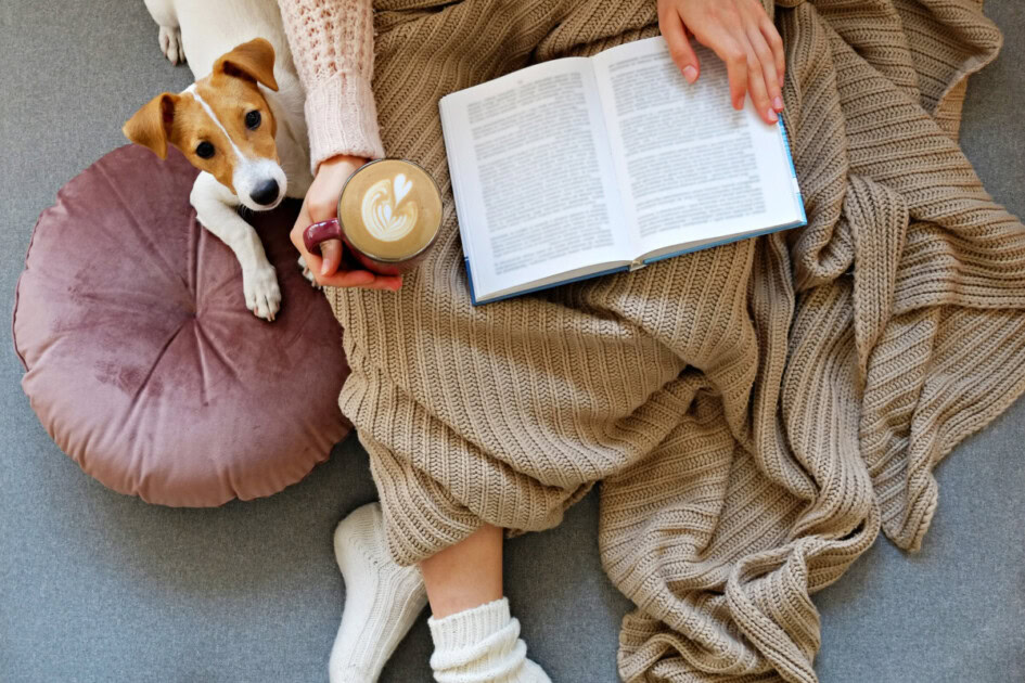 Woman holds coffee while reading with a blanket and Jack Russel Terrier next to her