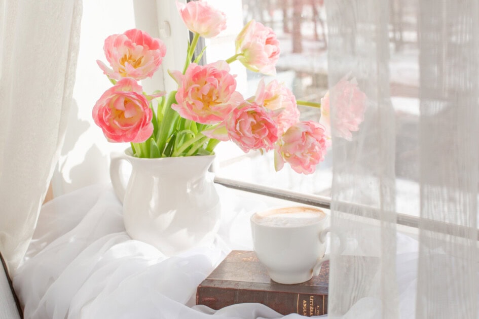 Bright pink flowers sit in vase next to window and mug of coffee