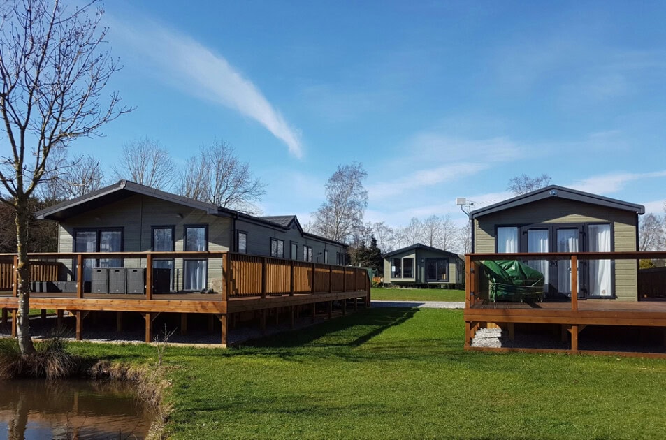 What a community might look like when buying a mobile home. A few green mobile homes with black trim sit on stained decks in a grassy field near a pond on a clear blue day. 