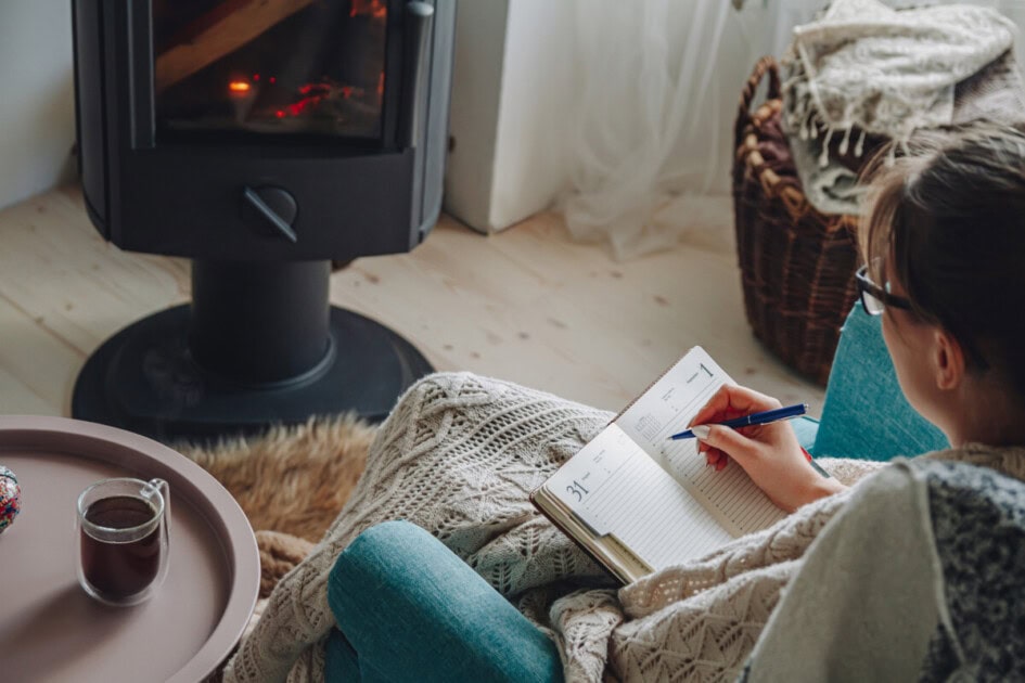 Woman writes in a journal with a blanket and hot drink in front of a fireplace