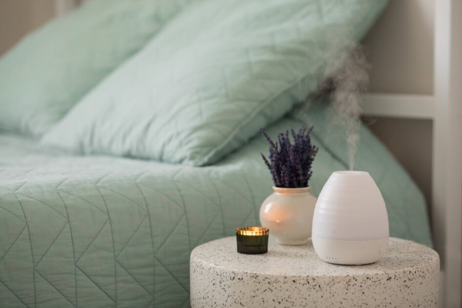 Oil diffuser and candle sit on table next to bed and lavender flowers